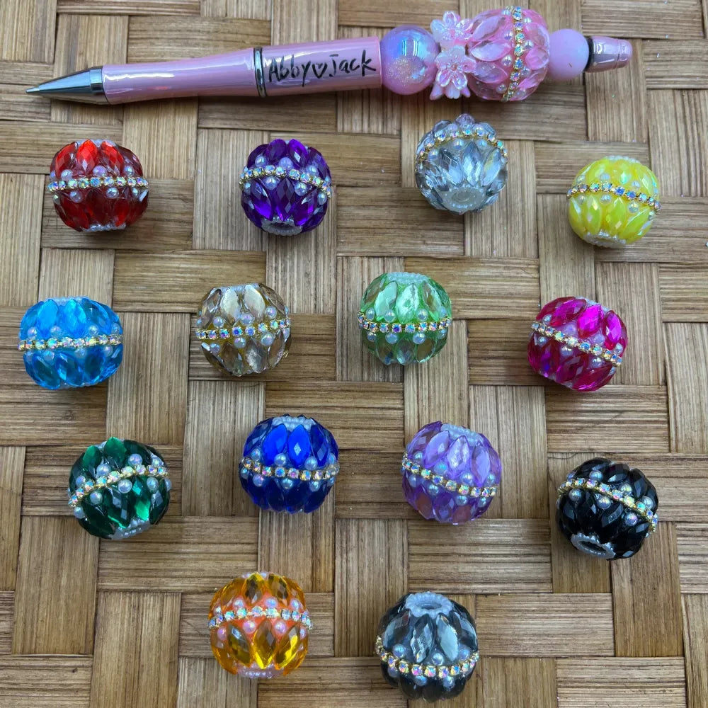 Assorted rhinestone and pearl-accented crystal faceted round fancy beads in 13 colors including red, purple, clear, yellow, blue, olive, green, pink, dark green, navy, lavender, black, orange, and gray displayed on a wicker surface next to a decorative beadable pen.