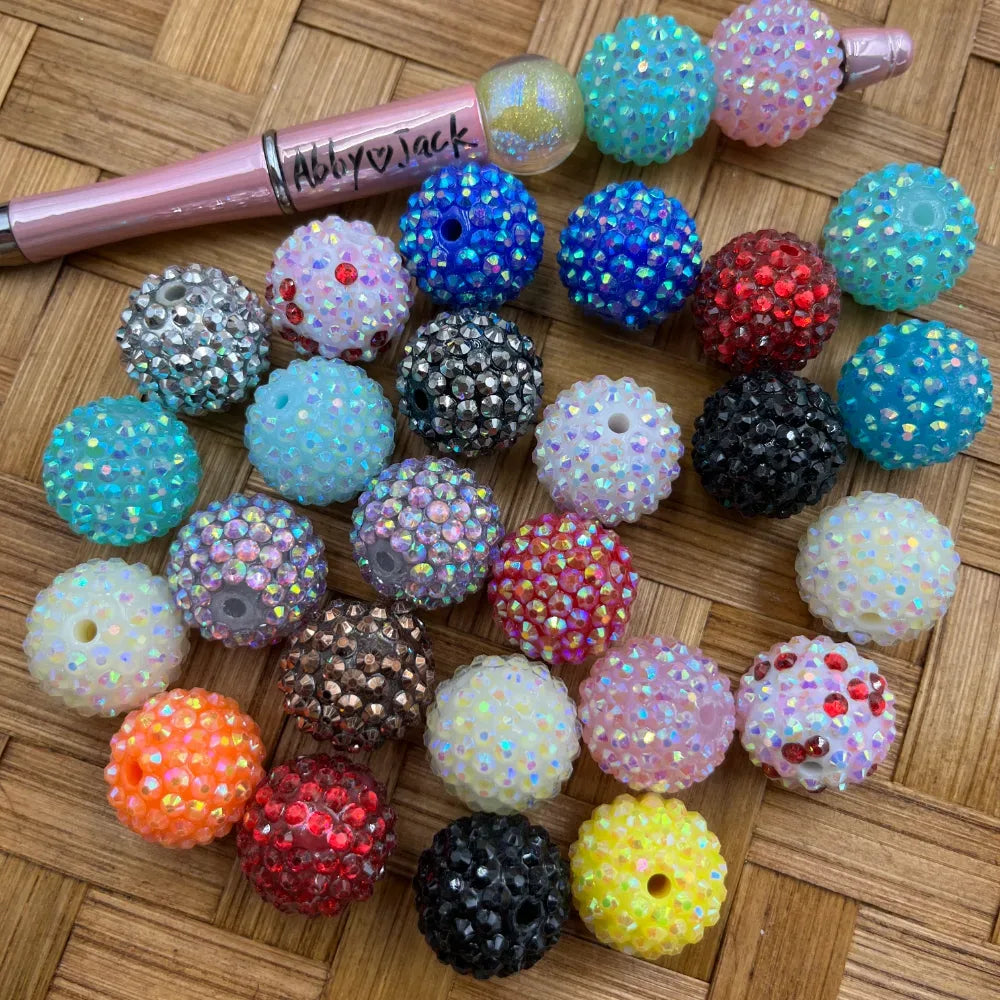 Assorted 20MM rhinestone sparkle acrylic beads in multiple colors including blue, teal, red, black, pink, orange, yellow, white, and gray on a woven bamboo surface next to a pink beadable pen branded Abby Jack.