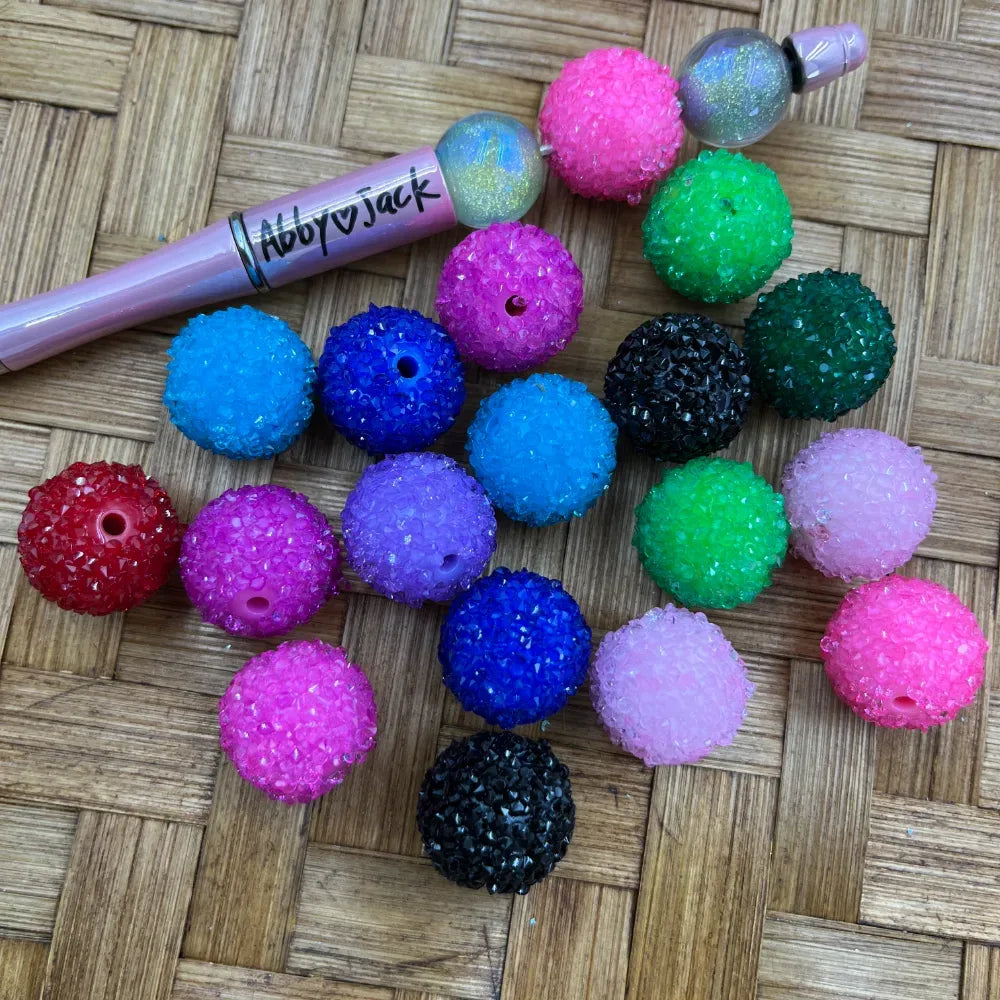 20MM colorful rhinestone sugar acrylic beads in pink, blue, green, purple, black and red displayed on a wicker surface next to a beadable pen from DIY Mini Planet.