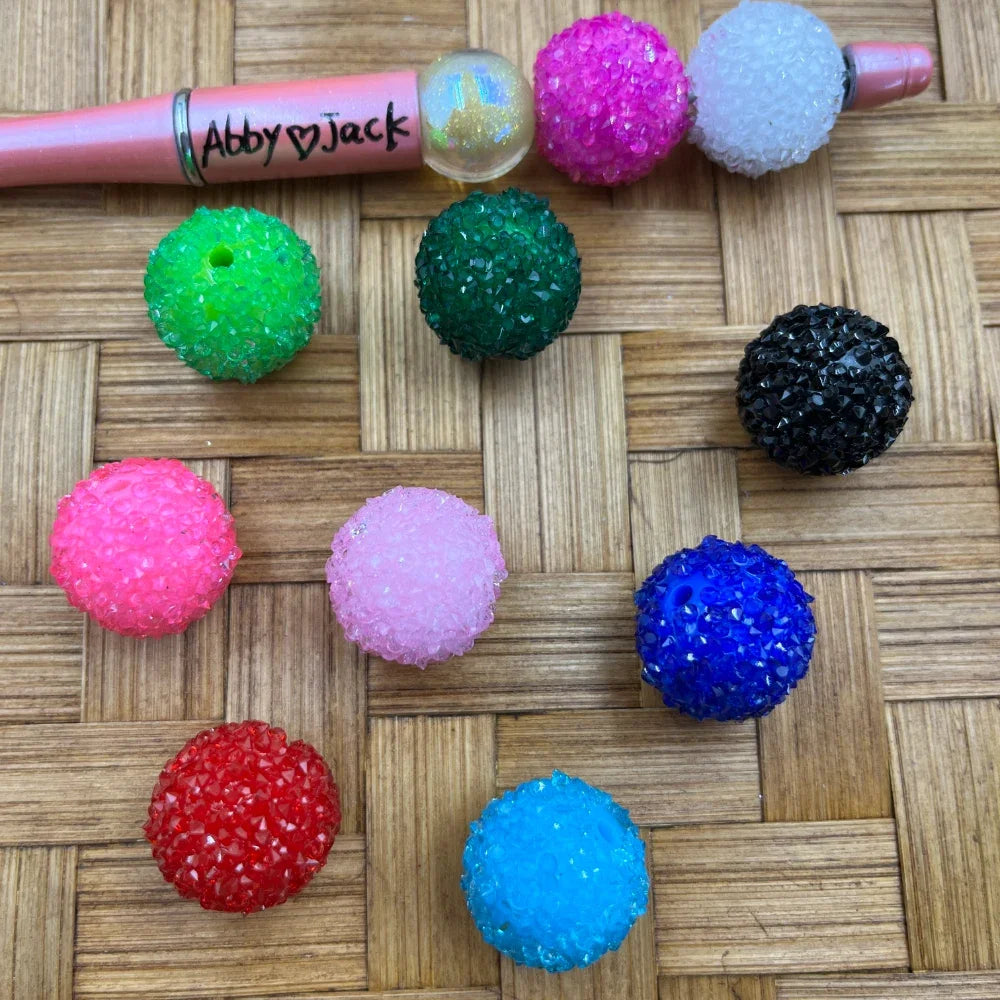 20MM rhinestone sugar beads in 10 colors including green, dark green, hot pink, white, black, blue, light blue, pink, red, and iridescent on a woven surface next to a pink beadable pen labeled Abby Jack.