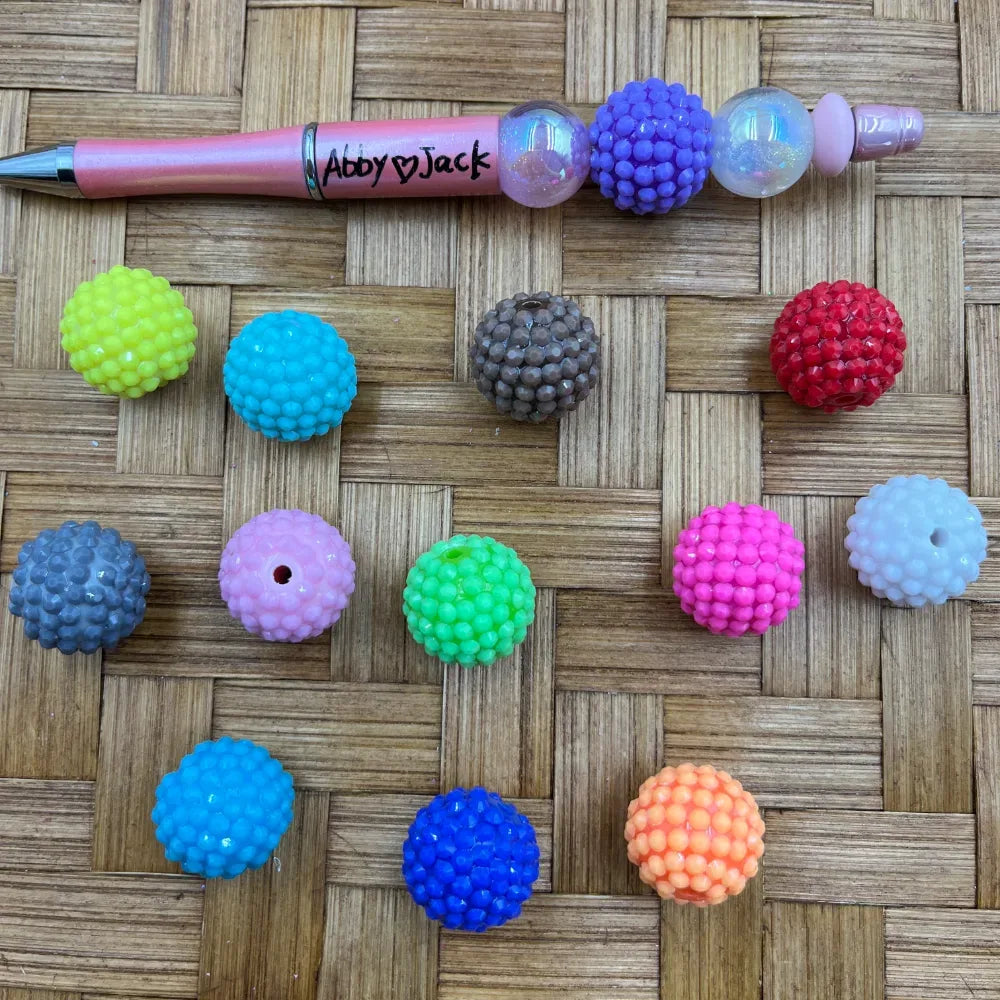 20MM solid color rhinestone beads in assorted colors including yellow, teal, pink, gray, blue, green, red, white, orange and more, displayed on a woven surface next to a beadable pen.