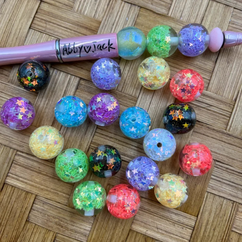 Colorful star confetti glitter acrylic beads in assorted colors including purple, green, red, black, blue, and yellow displayed on a wicker surface next to a pink beadable pen labeled Abby Jack.