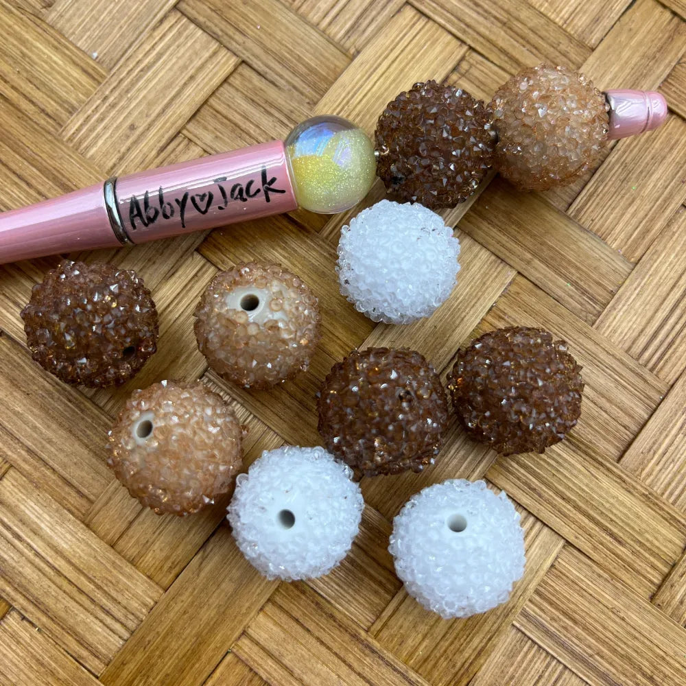 20MM sugar crystal rhinestone acrylic beads in brown, tan, and white scattered on wicker surface next to a pink beadable pen, showing textured bumpy rhinestone finish with center drill holes.