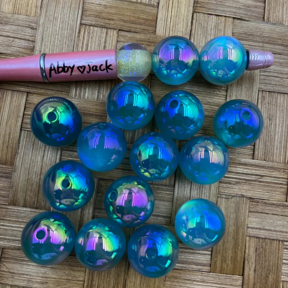 20MM iridescent teal aurora acrylic beads with holographic AB finish displayed next to a pink beadable pen on a wicker surface, showing blue-green-purple color shift.