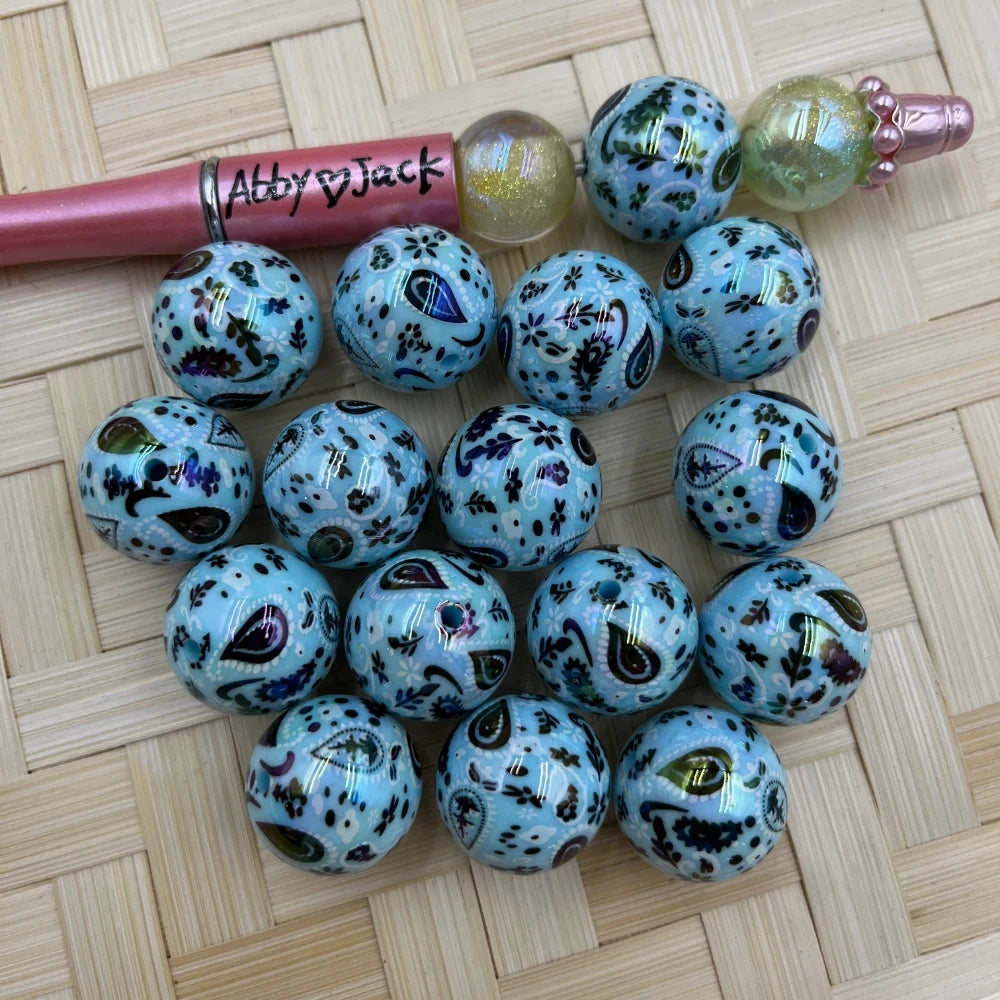 20MM teal paisley print acrylic beads with iridescent metallic finish arranged on a woven surface next to a beadable pen, showing detailed floral and teardrop paisley pattern in blue, purple, and green tones.