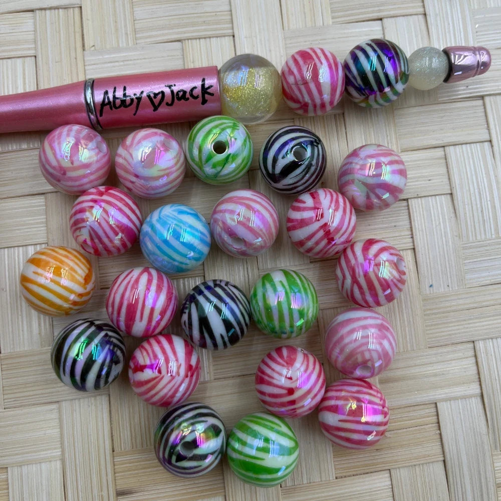20MM iridescent zebra stripe acrylic beads in assorted colors including pink, green, black, orange, and blue on a woven bamboo surface next to a pink beadable pen labeled Abby Jack.
