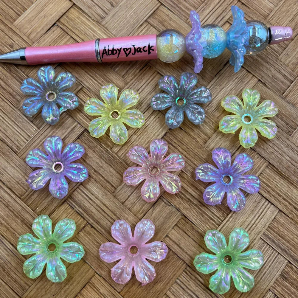 Assorted pastel iridescent daisy acrylic spacer beads with center holes beside a pink beadable pen
