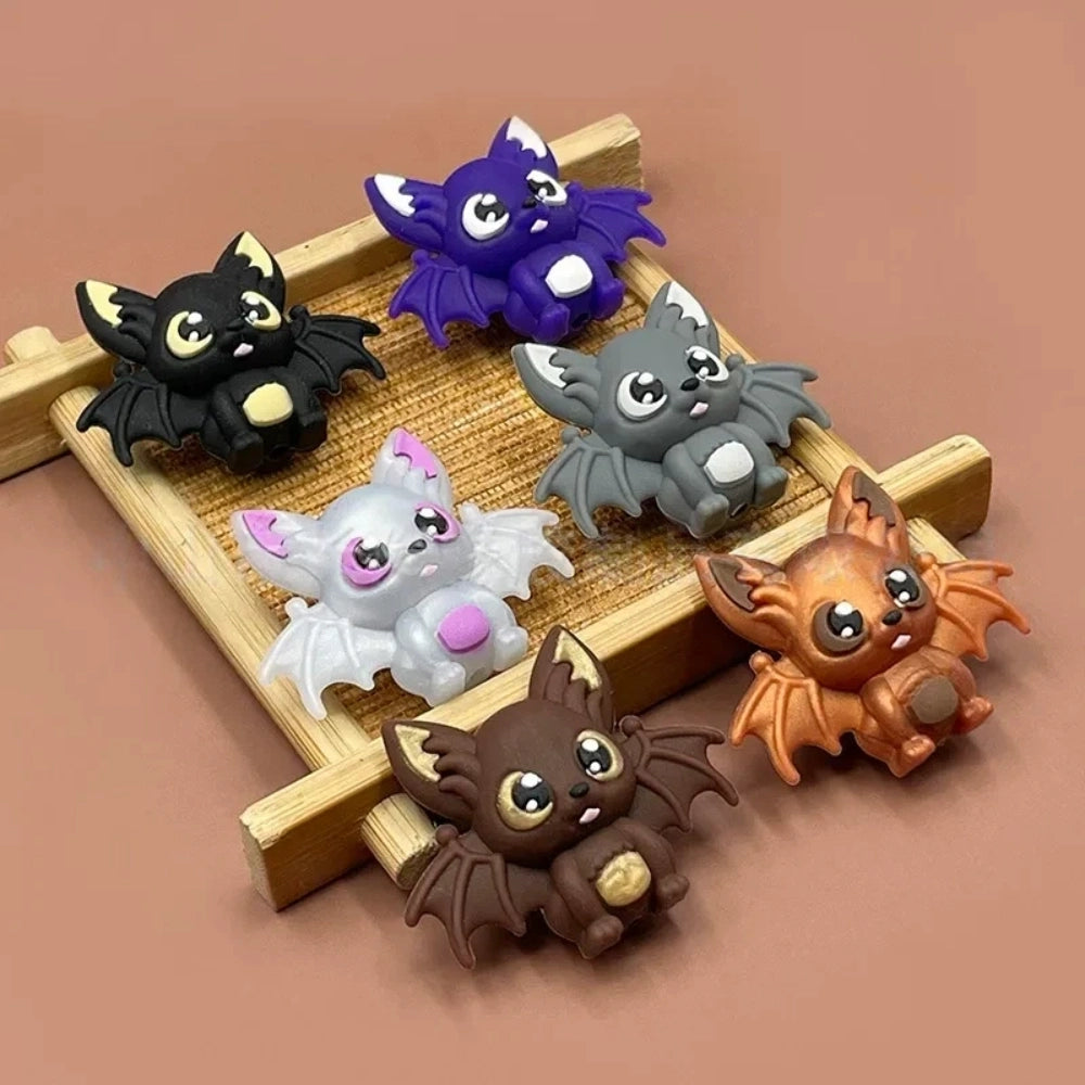 Six colorful 3D silicone baby bat focal beads in black, purple, gray, white, brown, and copper displayed on a wooden tray against a pink background.