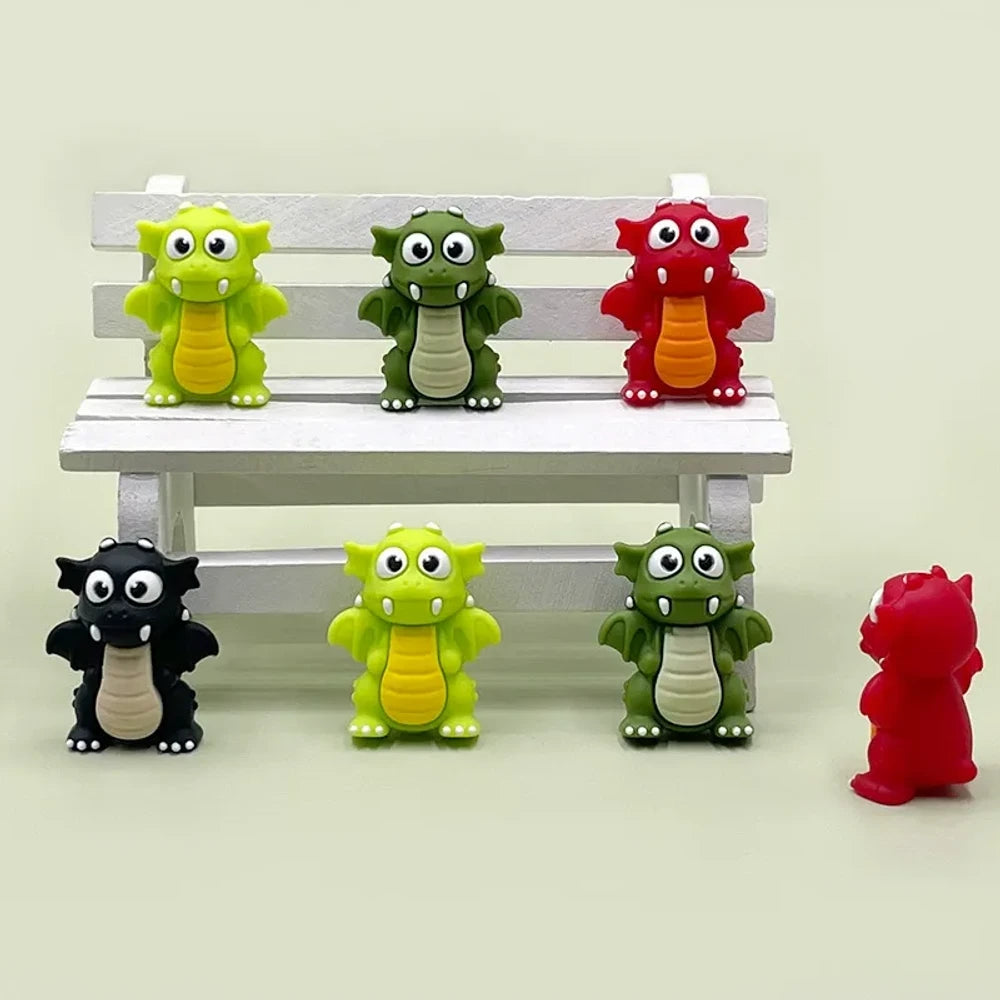Set of seven 3D cartoon dragon silicone focal beads in black, lime green, dark green, and red colors displayed on and around a miniature white wooden bench against a light green background.
