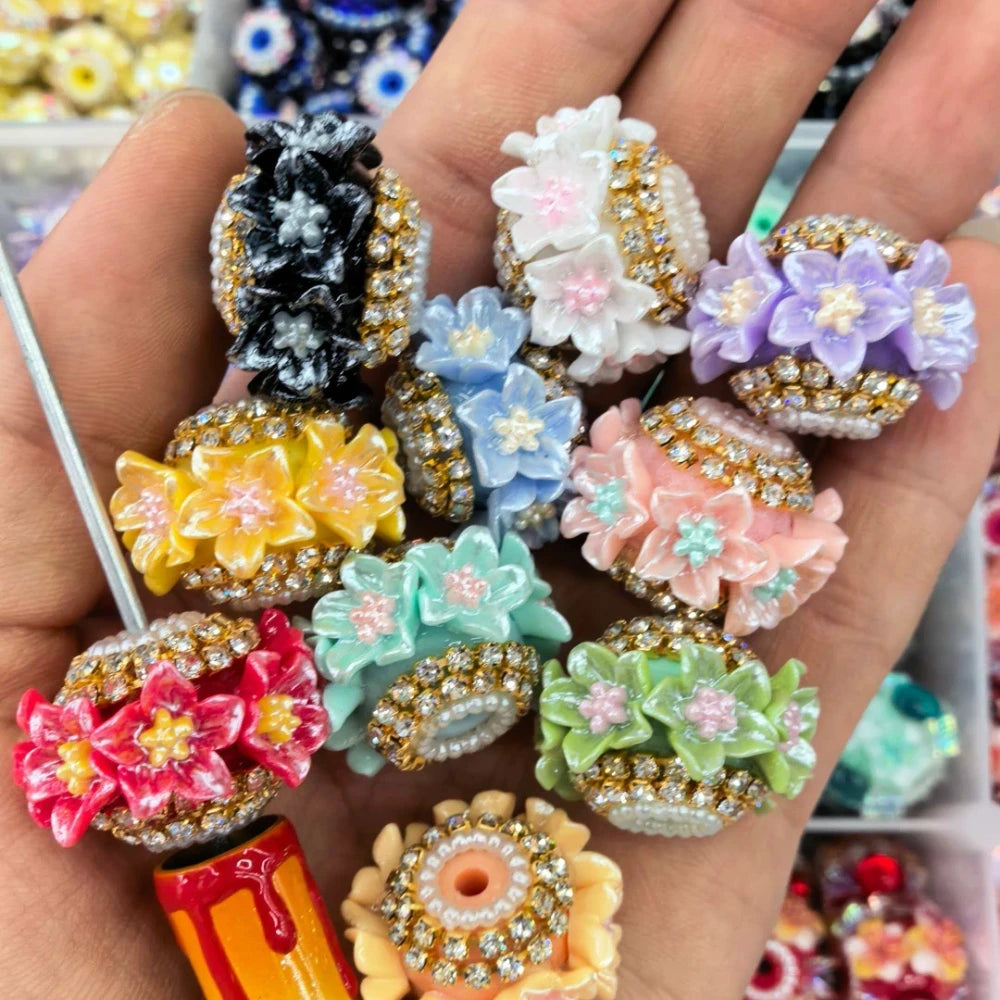 Assorted 3D flower rhinestone and pearl fancy beads in multiple colors including black, white, purple, yellow, blue, teal, pink, red, and green, held in a hand showing detailed resin floral designs with gold rhinestone and pearl borders.
