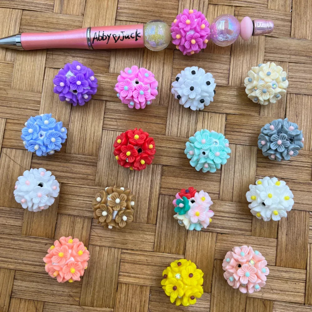 Assorted colorful 3D flower ball resin fancy beads in purple, pink, blue, red, teal, gray, white, yellow, peach, and multicolor, displayed on a woven rattan surface next to a beadable pen.