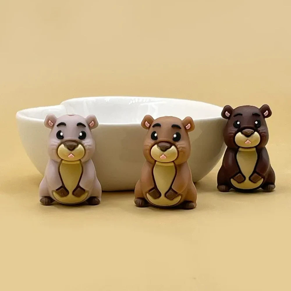 Three 3D groundhog silicone focal beads in light tan, medium brown, and dark brown colors displayed in front of a white ceramic bowl on a beige background.