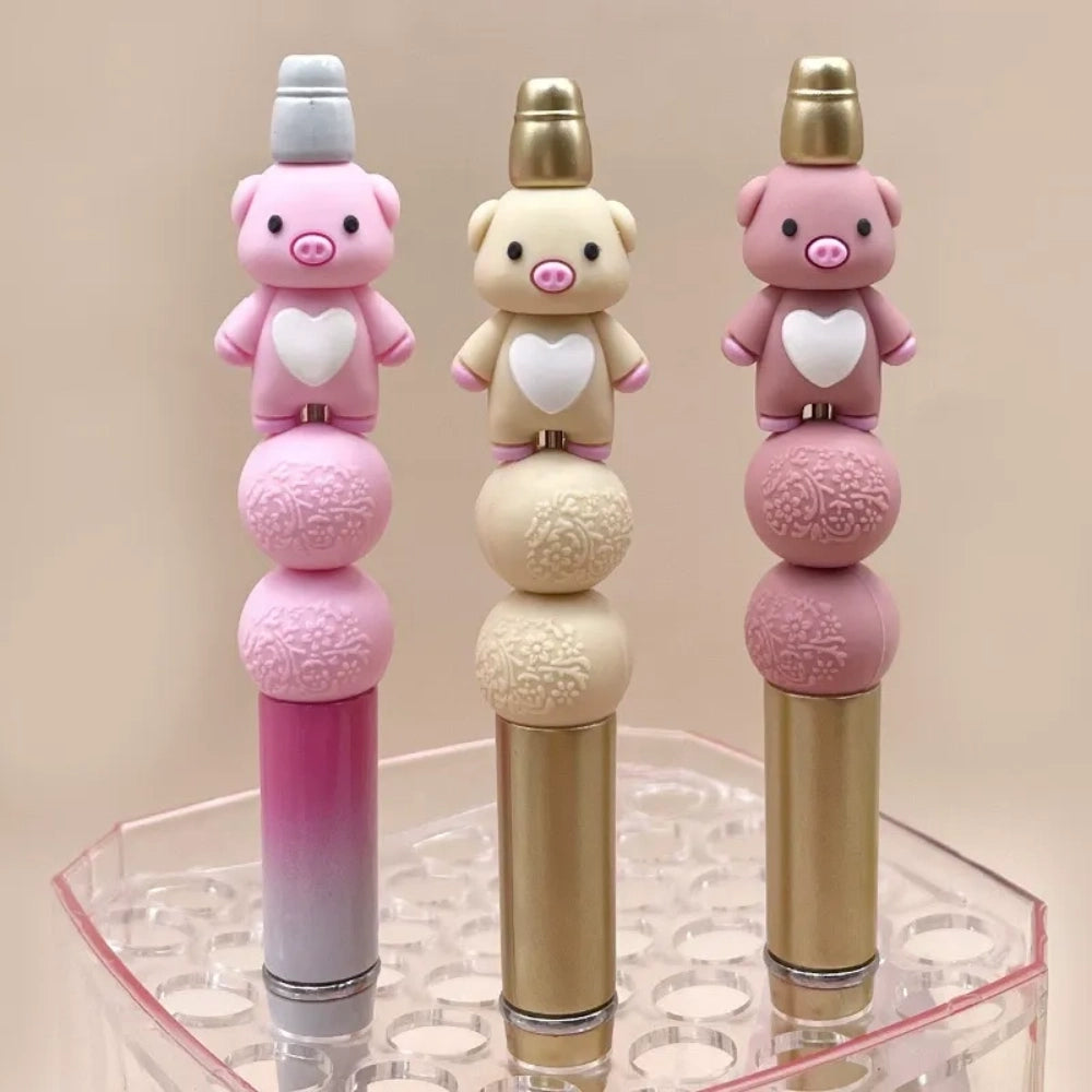 Three cute 3D pig silicone focal beads in pink, cream, and mauve colors, each featuring a white heart on the chest, displayed upright in a clear bead holder.