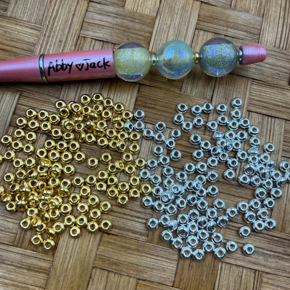 4MM gold and silver metallic metal spacer beads spread on a woven surface next to a pink beadable pen labeled Abby Jack, used for DIY jewelry making and crafts.