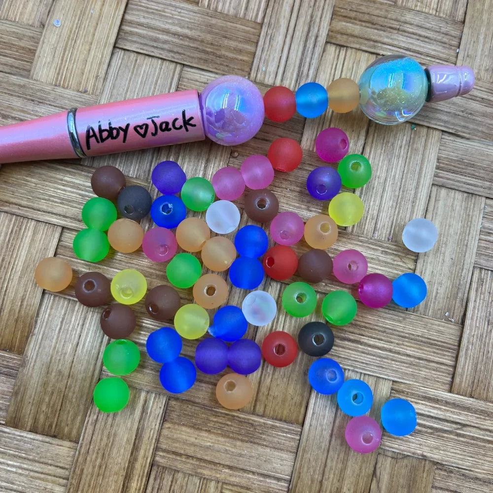 8MM frosted matte multicolor acrylic beads in assorted colors including blue, green, red, pink, yellow, purple, brown, and white, scattered next to a pink beadable pen on a wicker surface.