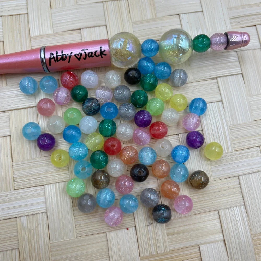 8MM mixed color glitter galaxy acrylic beads scattered on a wicker surface next to a pink beadable pen labeled Abby Jack, showing assorted translucent sparkle beads in blue, green, pink, purple, yellow, orange, and clear.