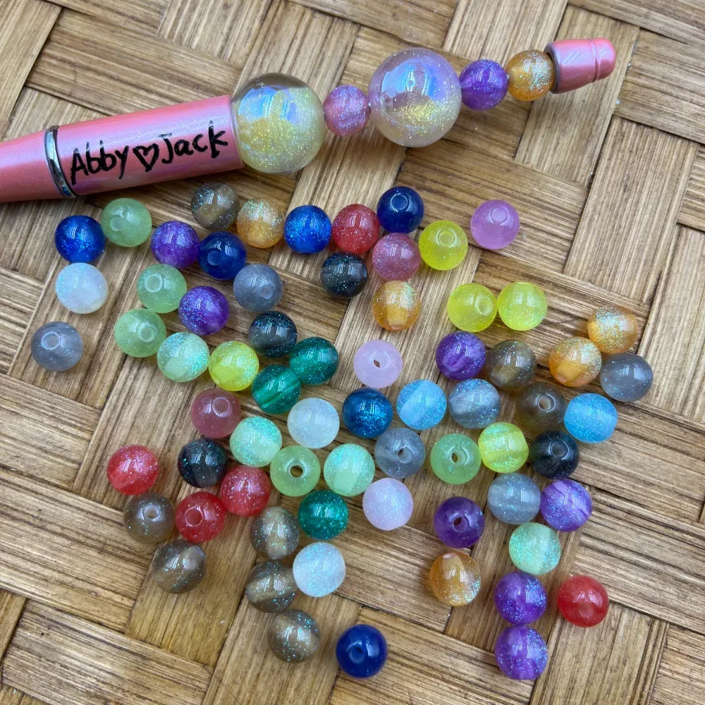 8MM glitter galaxy two-tone acrylic round beads in mixed colors including blue, purple, teal, yellow, red, pink, and gray with iridescent shimmer finish, displayed next to a beadable pen on a woven bamboo background.