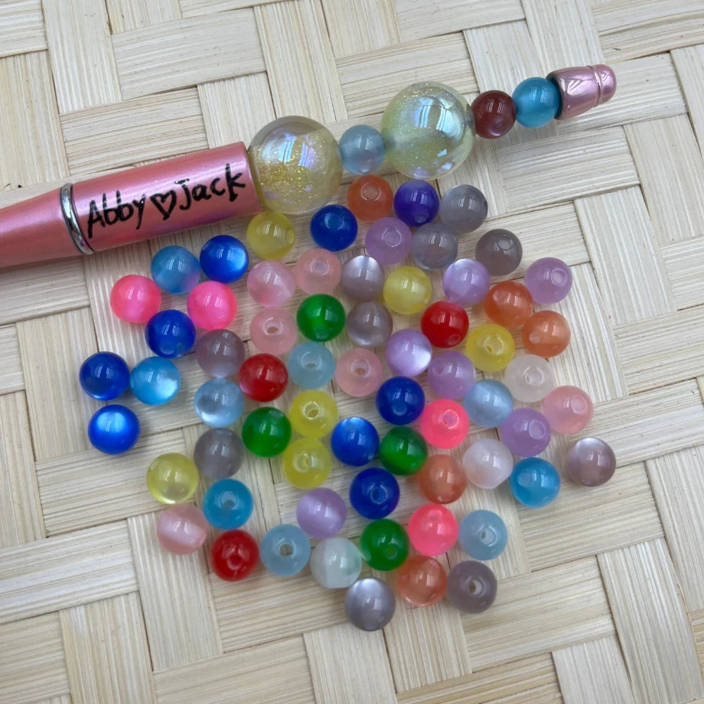 8MM mixed color glow moonstone acrylic beads scattered on a woven surface next to a pink beadable pen labeled Abby Jack, showing translucent and opaque round beads in assorted colors.