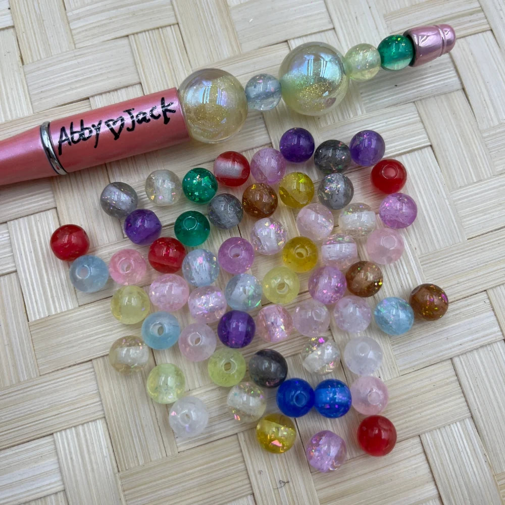 8MM iridescent glitter mix acrylic beads in assorted colors including pink, purple, teal, red, blue, yellow, and clear displayed next to a beadable pen on a woven surface.