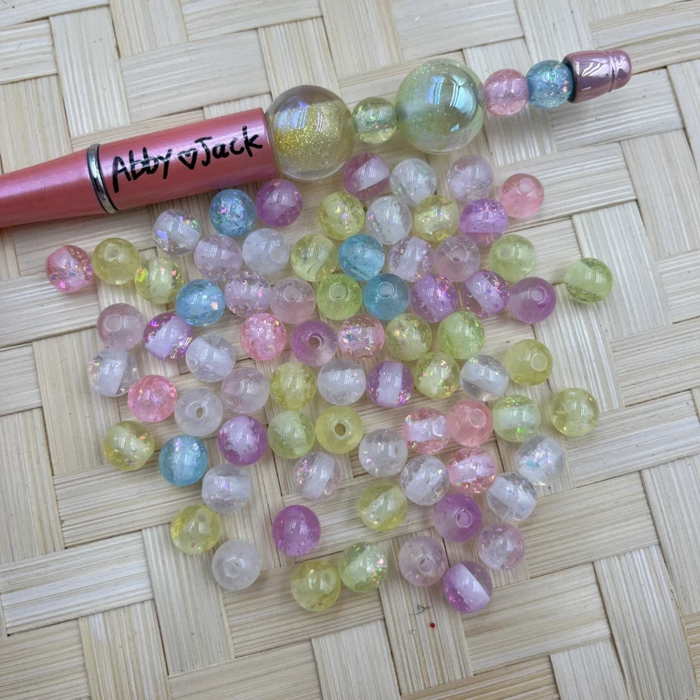 8MM pastel glitter confetti acrylic beads in pink, blue, yellow, purple, and white with iridescent flakes, shown next to a beadable pen on a woven surface.