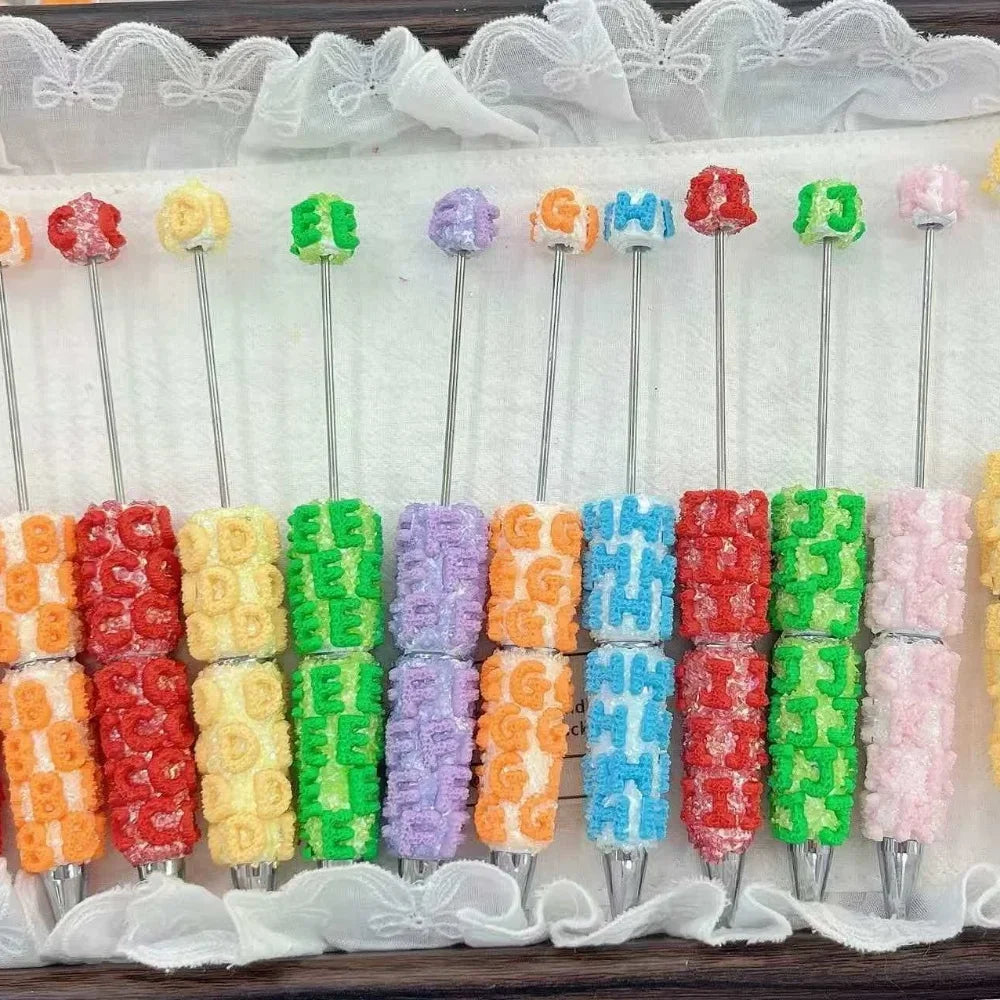 Colorful alphabet letter clay beadable pens A through J arranged in a row, each pen decorated with matching letter clay beads in red, orange, yellow, green, purple, orange, blue, red, green, and pink colors on a white lace fabric background.