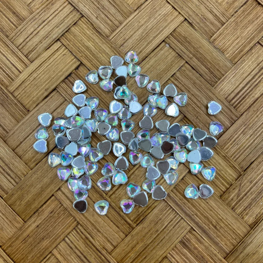 Small heart-shaped AB crystal flatback rhinestone fancy beads with iridescent rainbow shimmer and silver foil backing, scattered on woven bamboo surface, for DIY jewelry making and beadable pens.