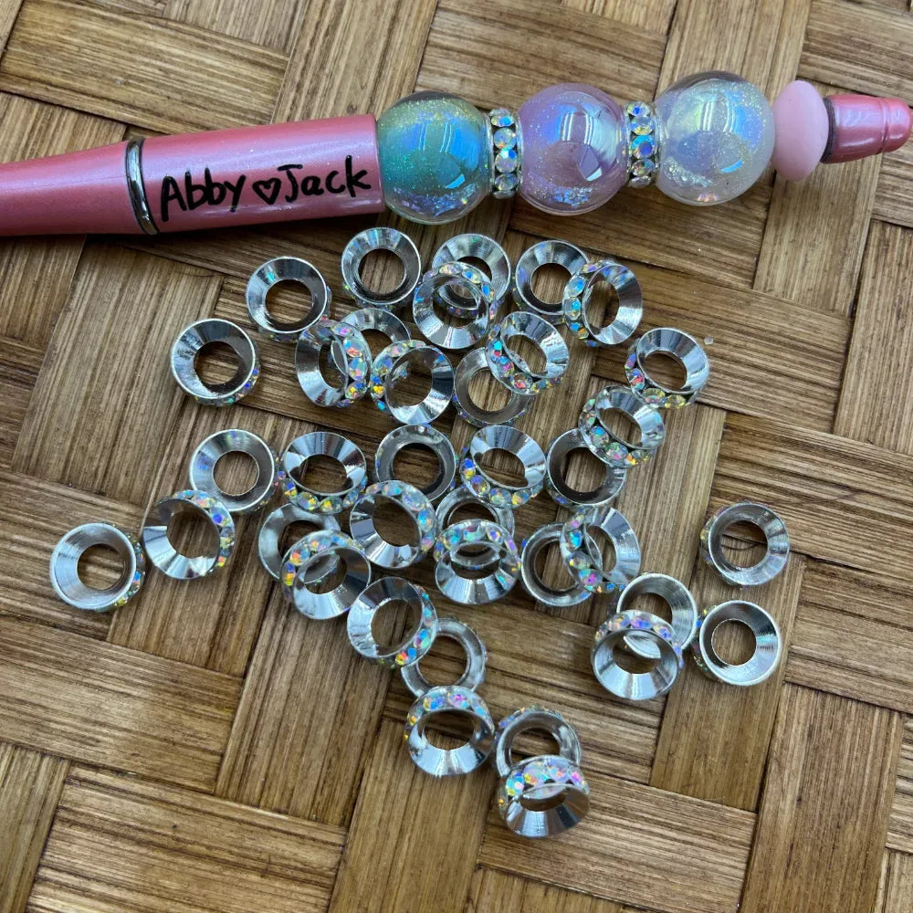 AB iridescent rhinestone silver metal ring spacer beads scattered on woven surface next to a pink beadable pen decorated with holographic globe beads and rhinestone spacers