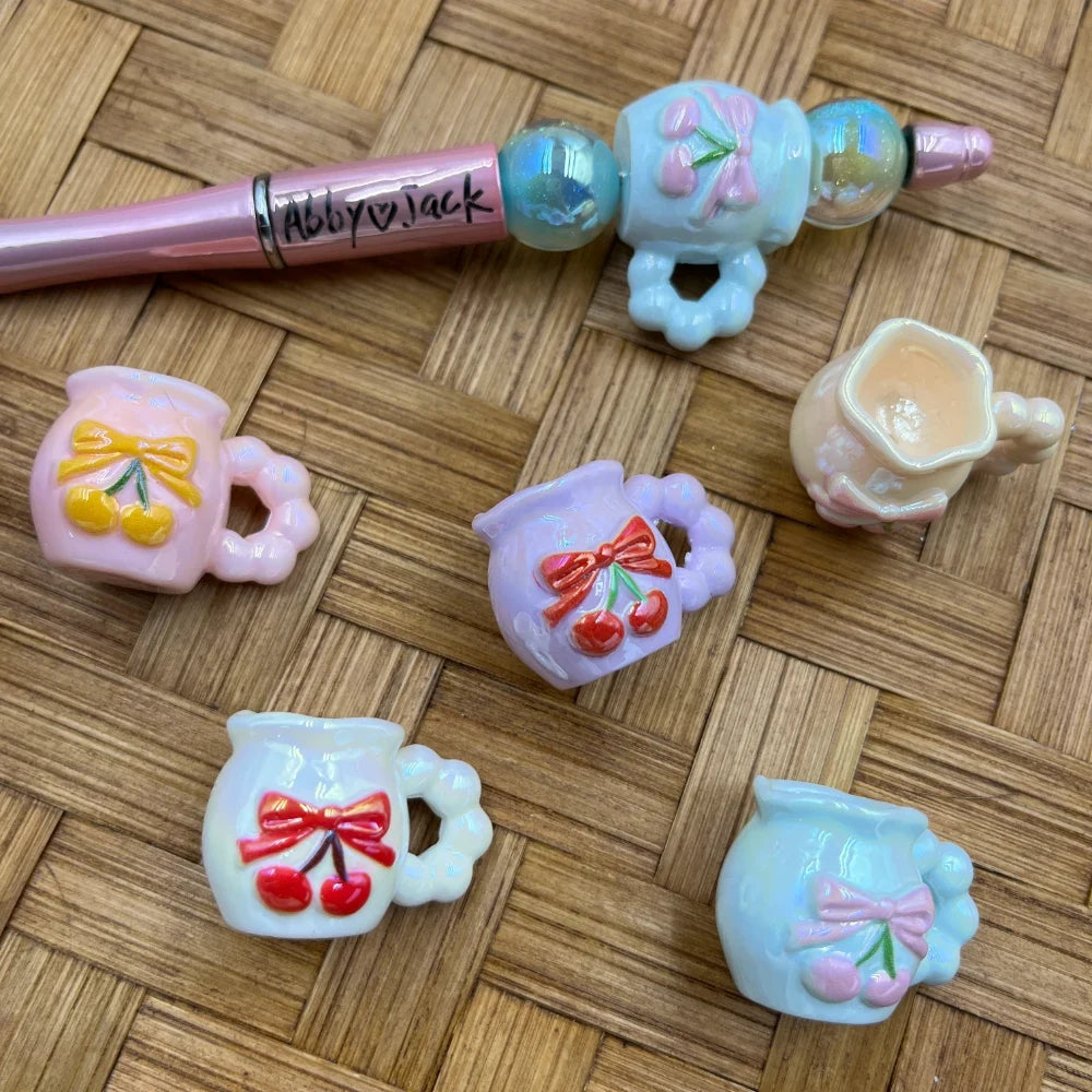 Assorted pastel cherry bow mug acrylic beads in pink, lavender, white, blue, and cream with red bow and cherry designs, shown with a beadable pen on wicker background.