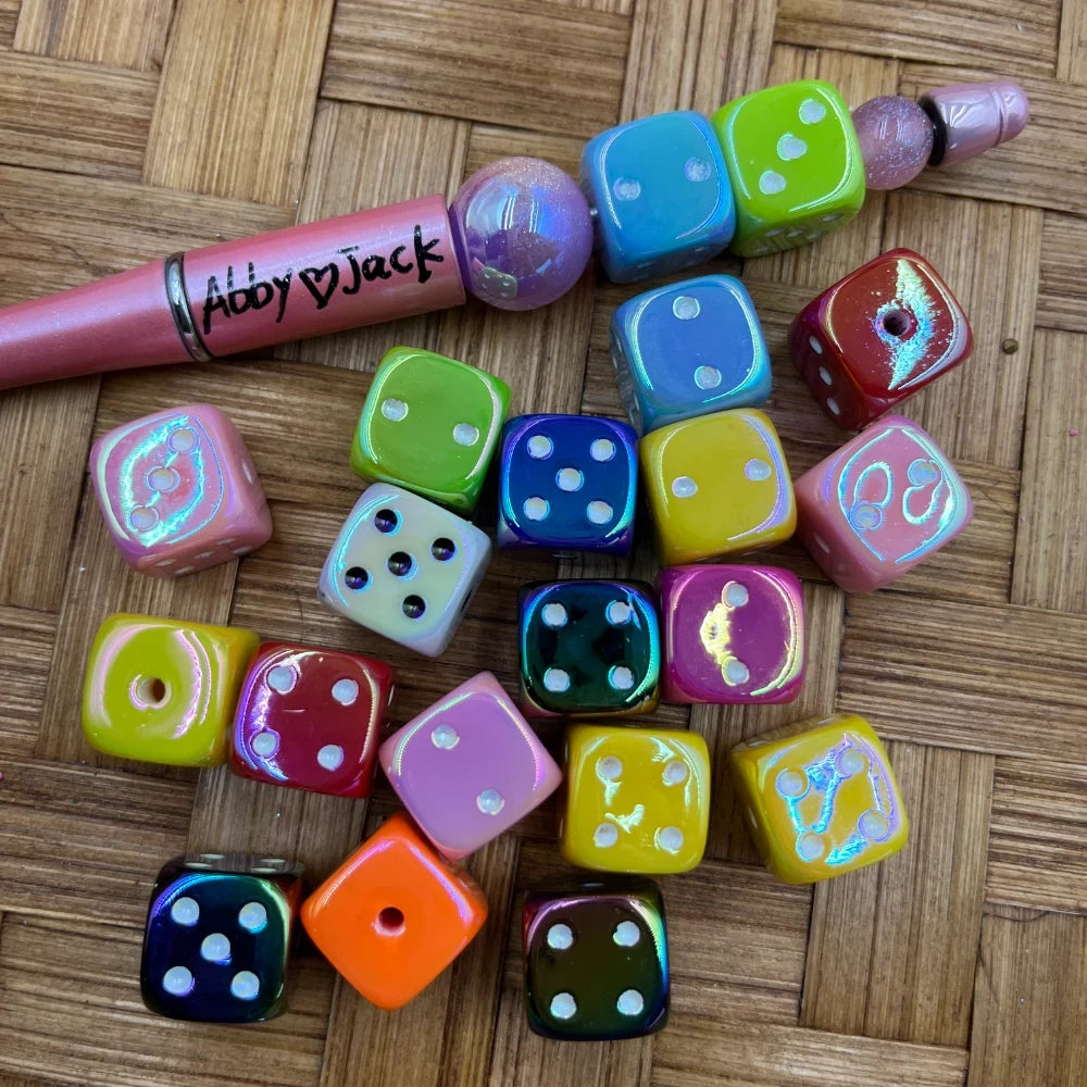 Assorted iridescent dice-shaped acrylic beads in mixed colors including pink, blue, green, yellow, orange, red, and black, displayed on a wicker surface next to a beadable pen labeled Abby and Jack.