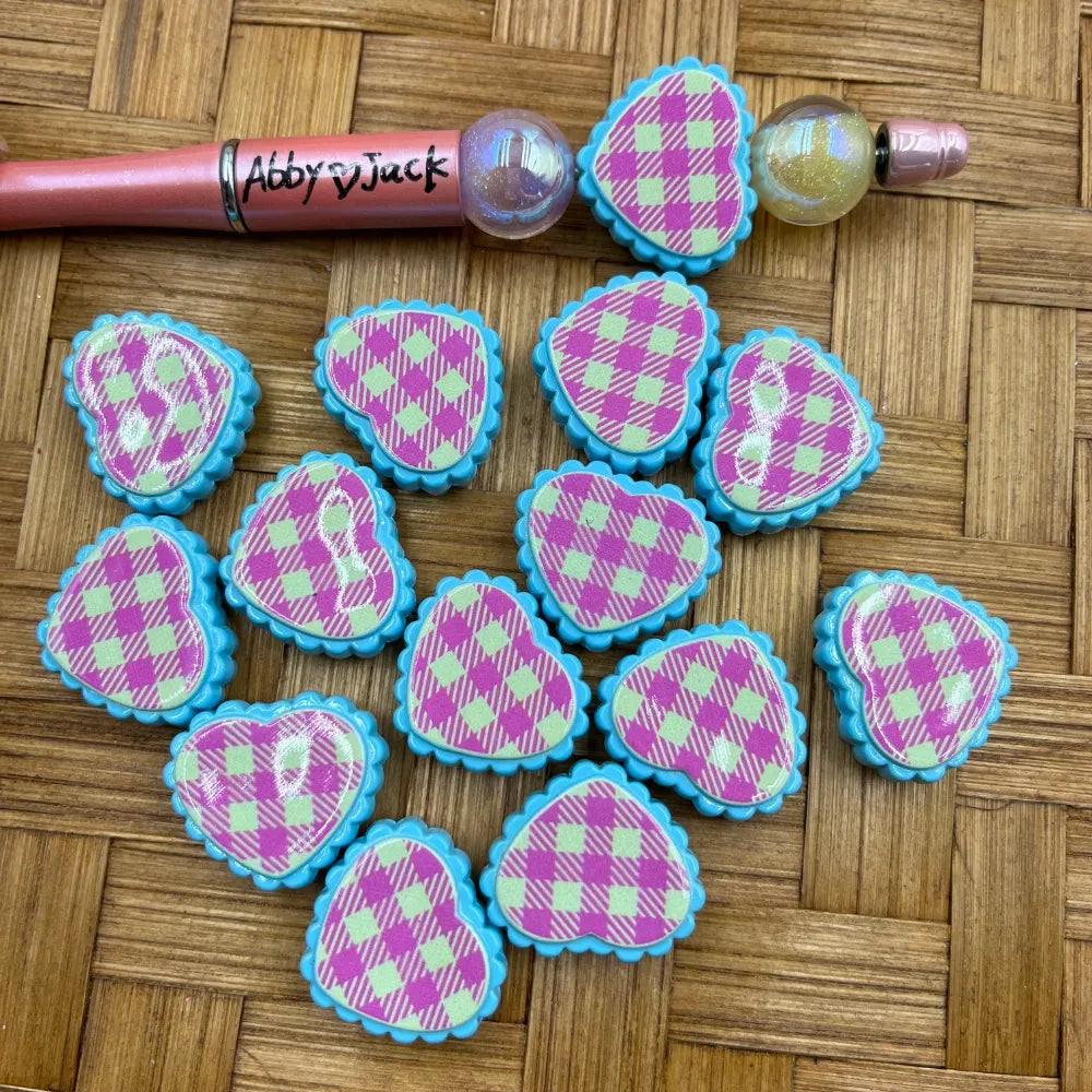 Pink and green gingham plaid heart-shaped acrylic beads with teal scalloped edges arranged on a wicker surface next to beadable pens