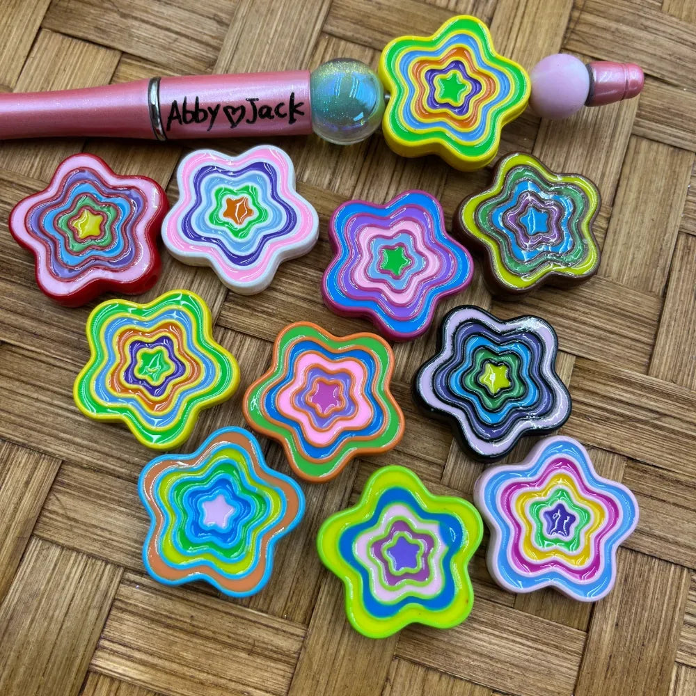 Colorful layered rainbow star flower acrylic beads in multiple color combinations displayed on a woven surface next to a beadable pen, showing concentric star flower ring patterns in neon, pastel, and dark colorways.
