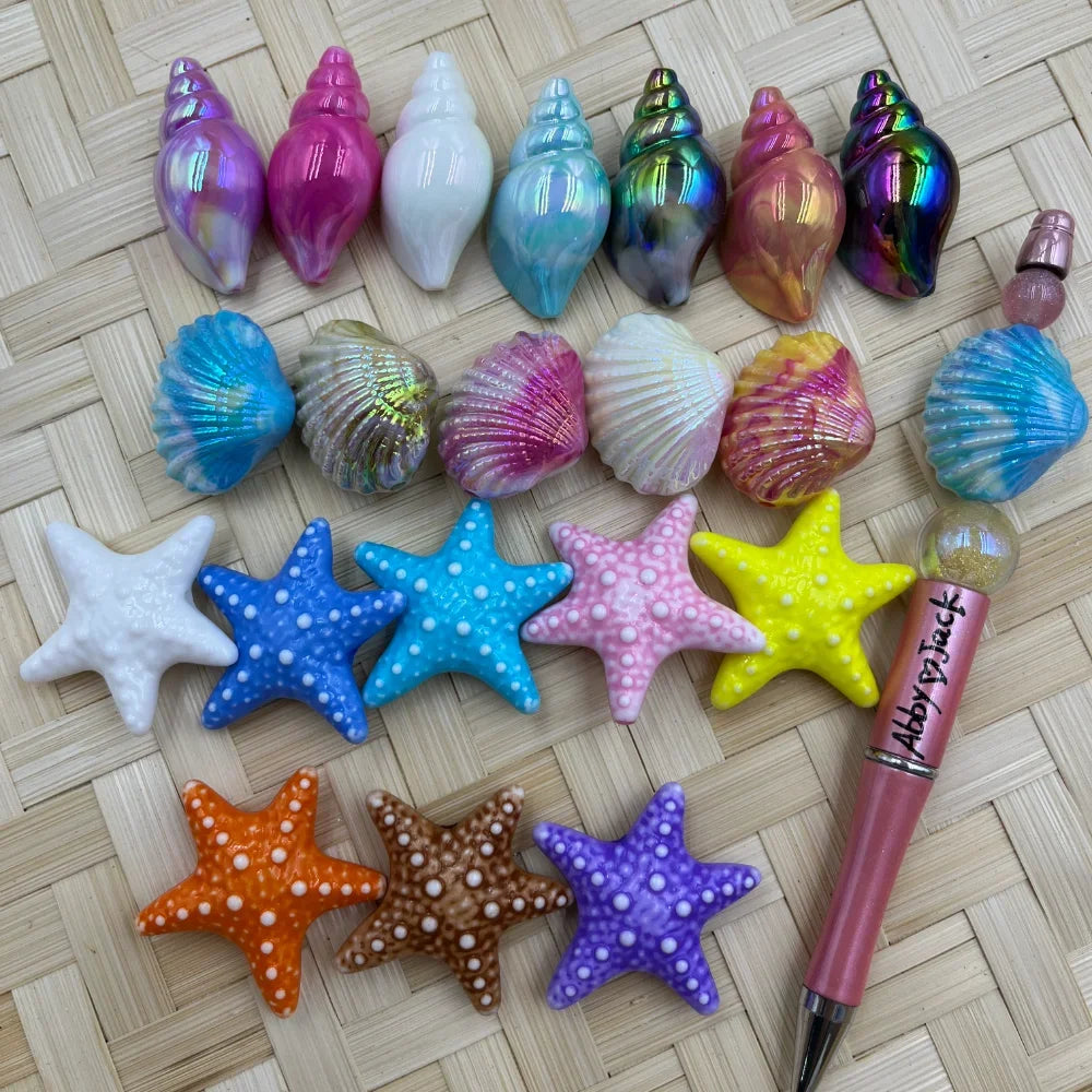 Assorted colorful acrylic ocean-themed beads including conch shells, scallop shells, and starfish in iridescent, pearlescent, and vibrant colors displayed on a woven surface next to a pink beadable pen.