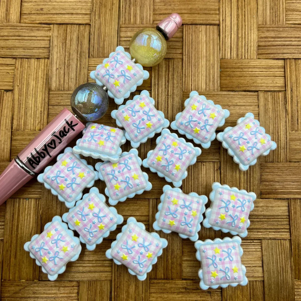 Flat square acrylic beads with pink gingham pattern, blue bow details, yellow stars, and scalloped white edges arranged on a wicker surface next to a pink pen and glitter ball beads.