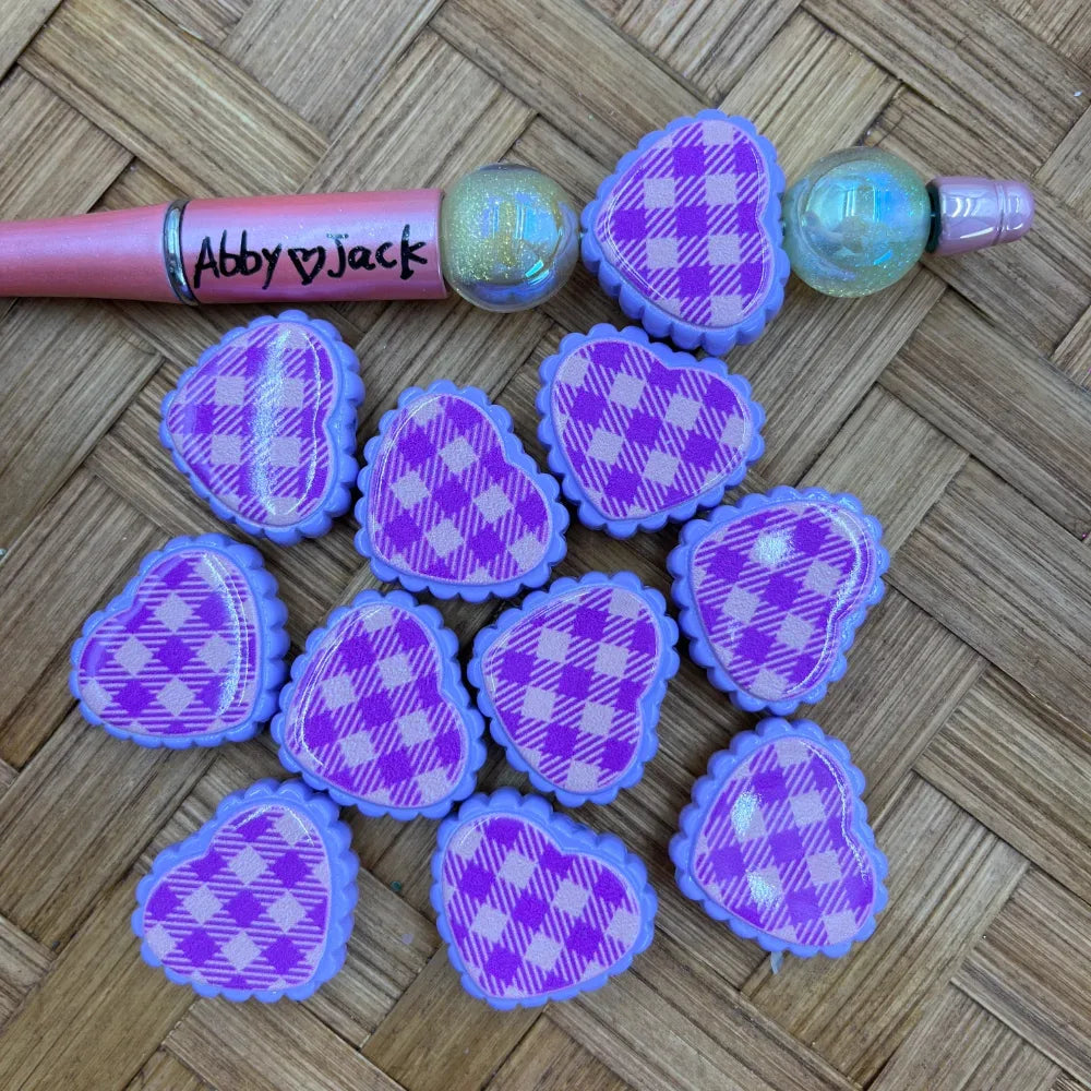 Purple and lavender plaid pattern Easter egg shaped acrylic beads with scalloped edges displayed on wicker surface next to a beadable pen