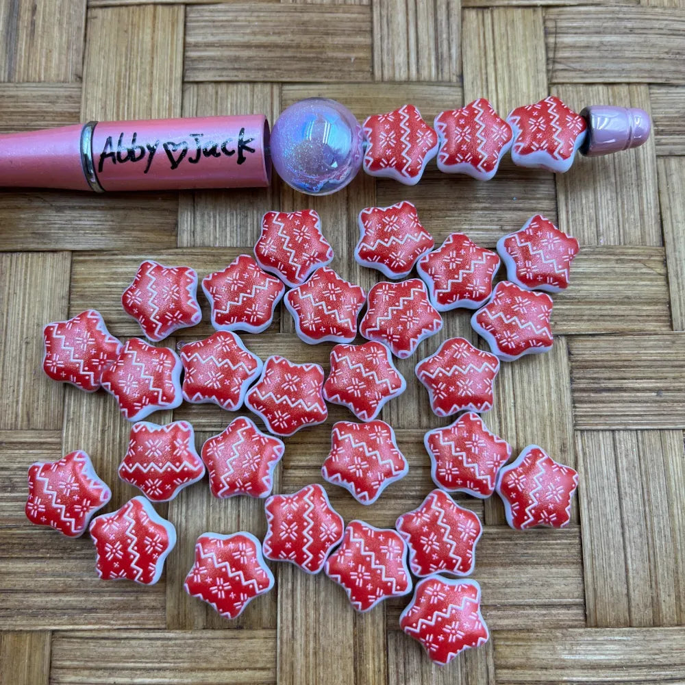 Red and white Nordic Fair Isle sweater-shaped acrylic beads displayed next to a pink beadable pen on a woven bamboo surface