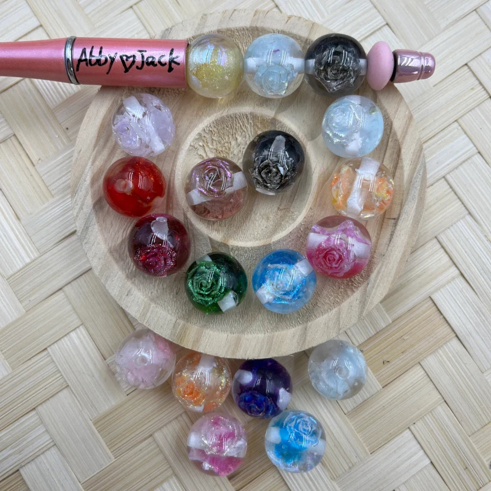 Assorted colorful acrylic rose flower glitter beads in multiple colors including red, blue, green, pink, black, and orange, displayed on a wooden tray with a beadable pen, showing embedded rose designs with glitter finish.
