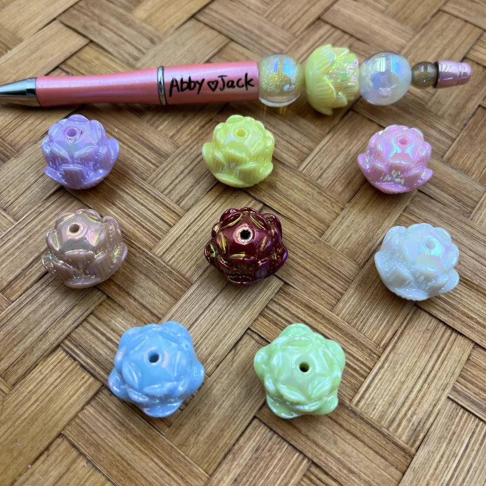 Assorted iridescent AB finish rose flower acrylic beads in pastel purple, yellow, pink, white, blue, green, mauve, and dark red, displayed on woven bamboo surface next to a beadable pen.