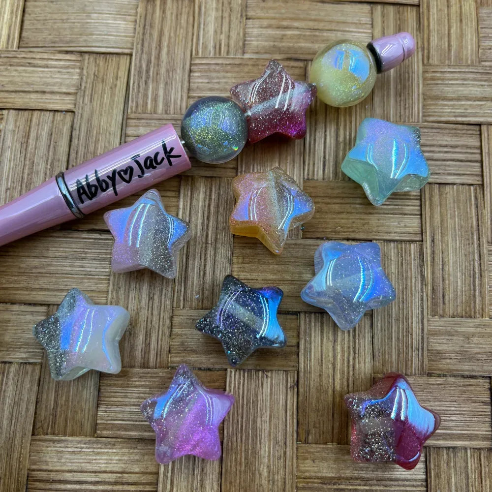Assorted iridescent galaxy star-shaped acrylic beads with glitter shimmer and aurora finish in multiple colors including pink, blue, black, orange, and clear, displayed on a wicker surface next to a pink beadable pen.