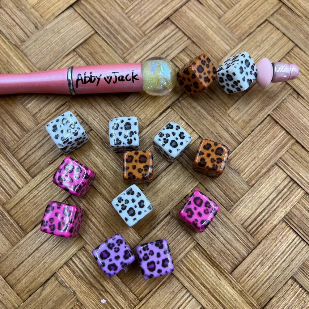 Assorted leopard print acrylic cube beads in pink, orange, white, and purple with black spots, shown with a beadable pen on a wicker surface