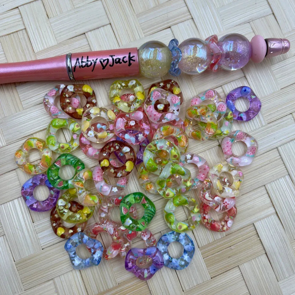 Colorful fruit and floral patterned acrylic donut spacer beads in assorted colors including pink, green, purple, and brown, displayed next to a beadable pen on a wicker surface.
