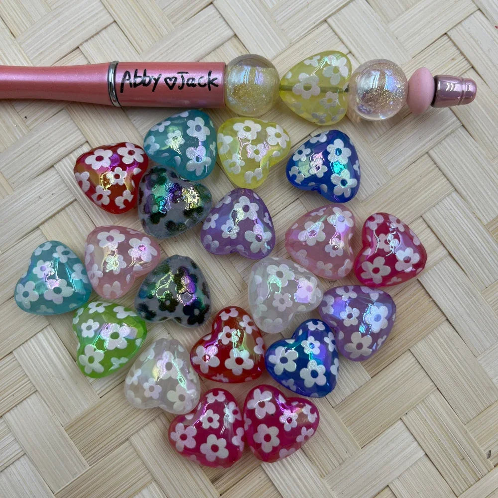 Colorful heart-shaped acrylic beads with white daisy floral print in assorted colors including red, blue, green, purple, pink, and yellow, displayed on a wicker surface next to a beadable pen.