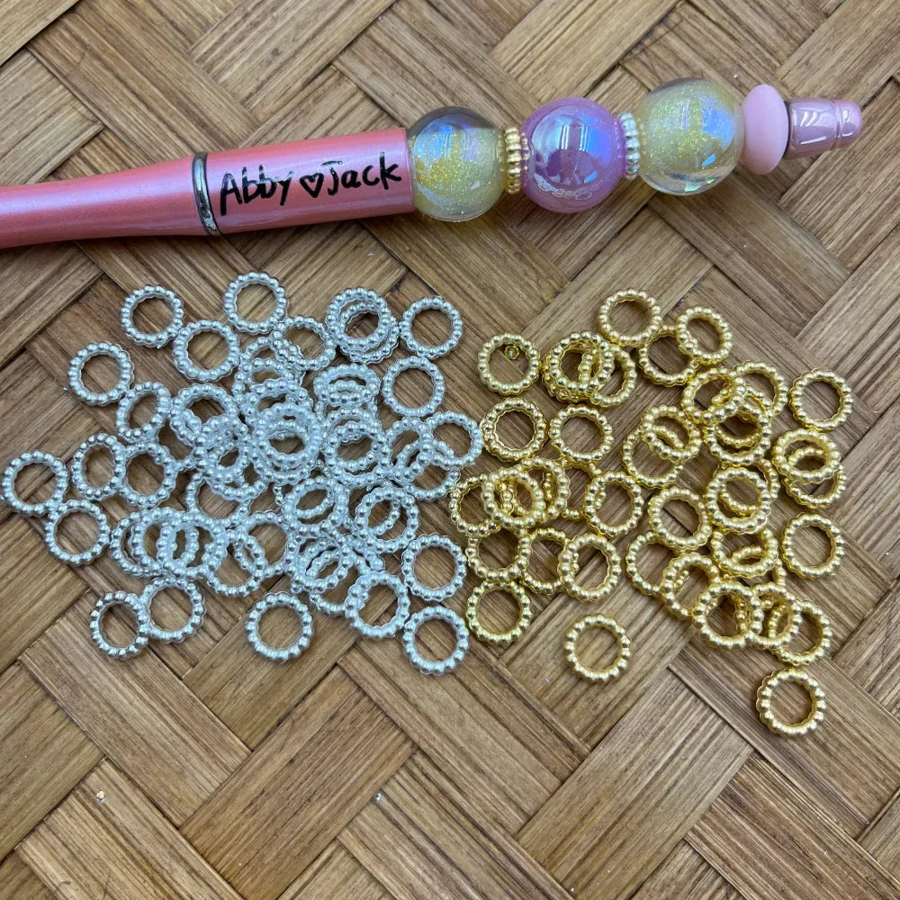 Silver and gold pearl beaded ring acrylic spacer beads displayed on a woven surface next to a decorated beadable pen, showing circular ring shape with beaded texture edges.