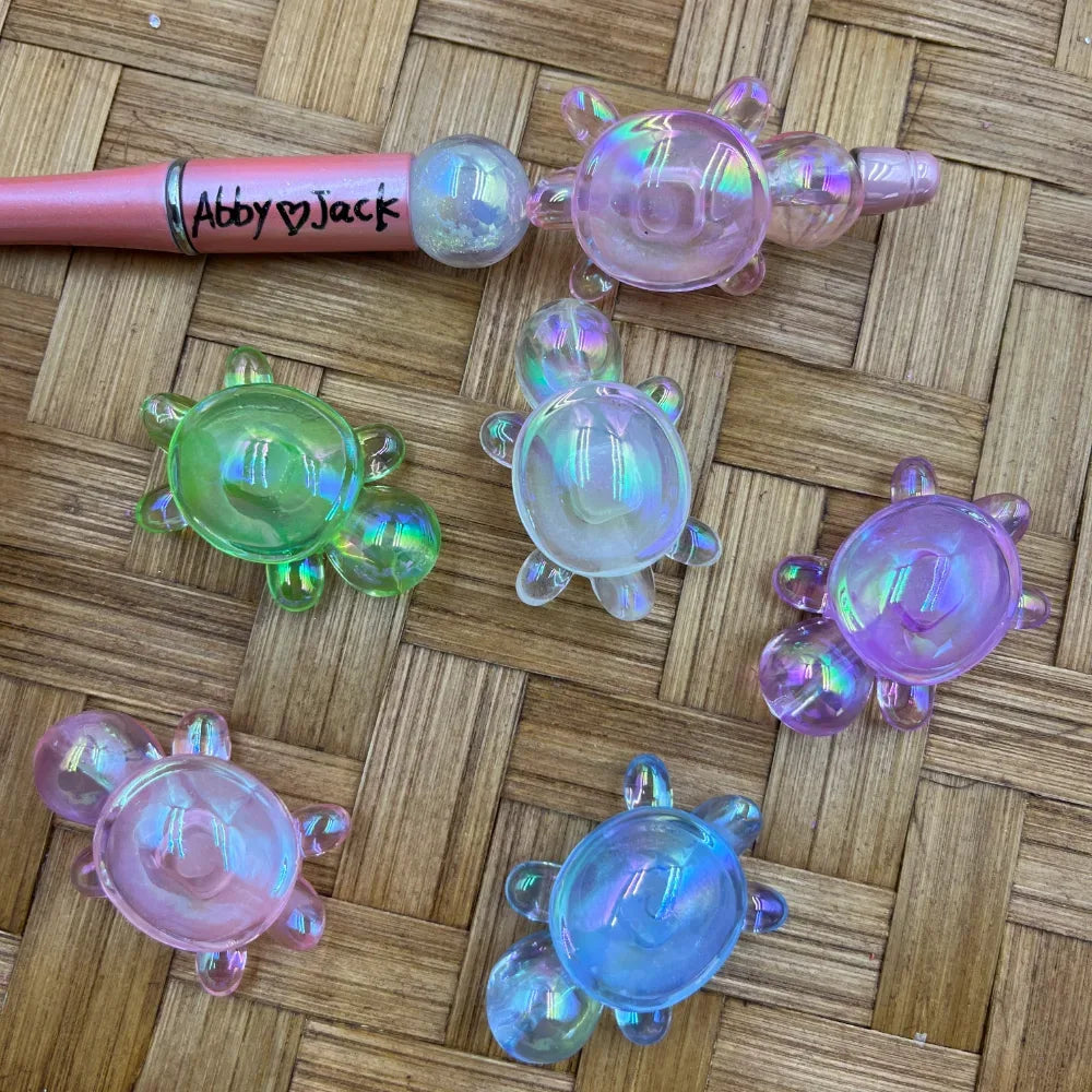Iridescent pastel acrylic turtle beads in green, pink, purple, blue, and clear rainbow colors displayed on wicker surface next to a pink beadable pen labeled Abby Jack.