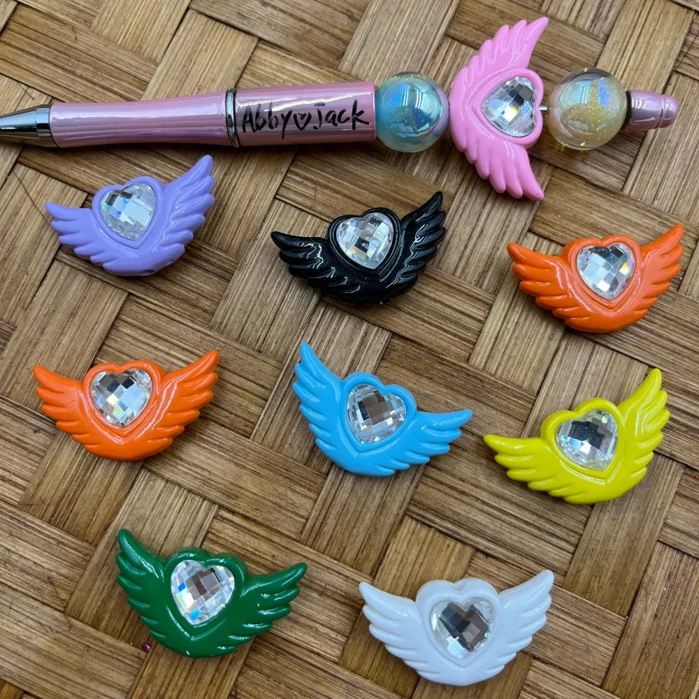 Colorful angel wing heart crystal acrylic beads in purple, black, orange, blue, yellow, green, white, and pink displayed on wicker surface with a beadable pen.