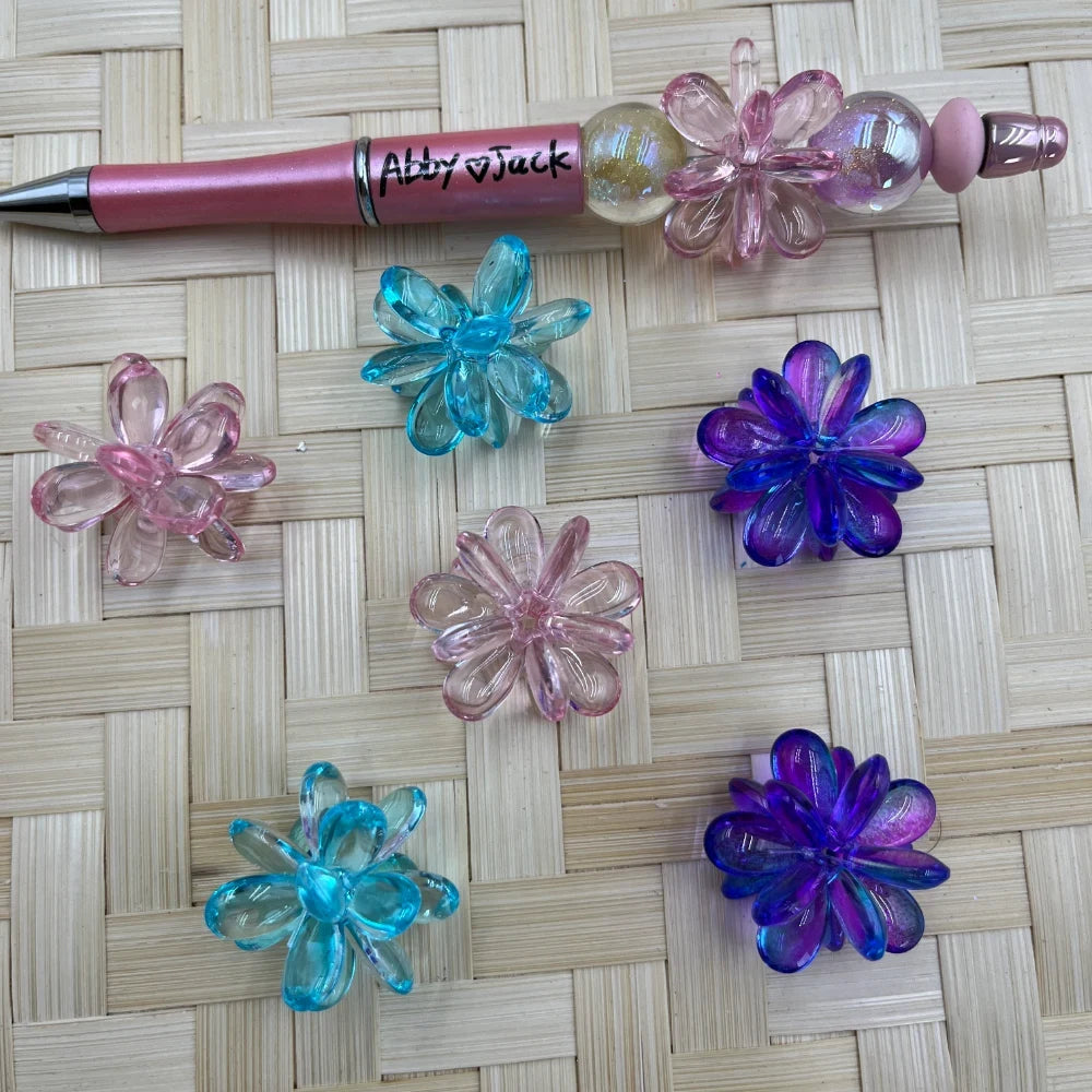 Assorted translucent iridescent acrylic flower beads in pink, aqua blue, and purple beside a pink beadable pen.