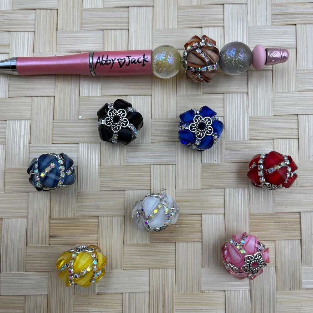 Assorted swirl acrylic fancy beads with rhinestone bands and silver-tone accents beside a pink beadable pen
