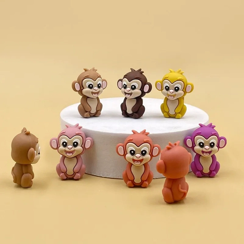 Set of 7 colorful baby monkey 3D silicone focal beads in brown, dark brown, yellow, pink, orange, rust, and purple displayed on a white pedestal with yellow background.