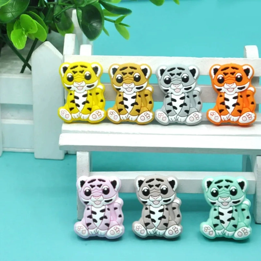 Seven colorful baby tiger silicone focal beads in yellow, brown, gray, orange, pink, white, and mint displayed on a miniature white bench against a teal background. - Random Mixed