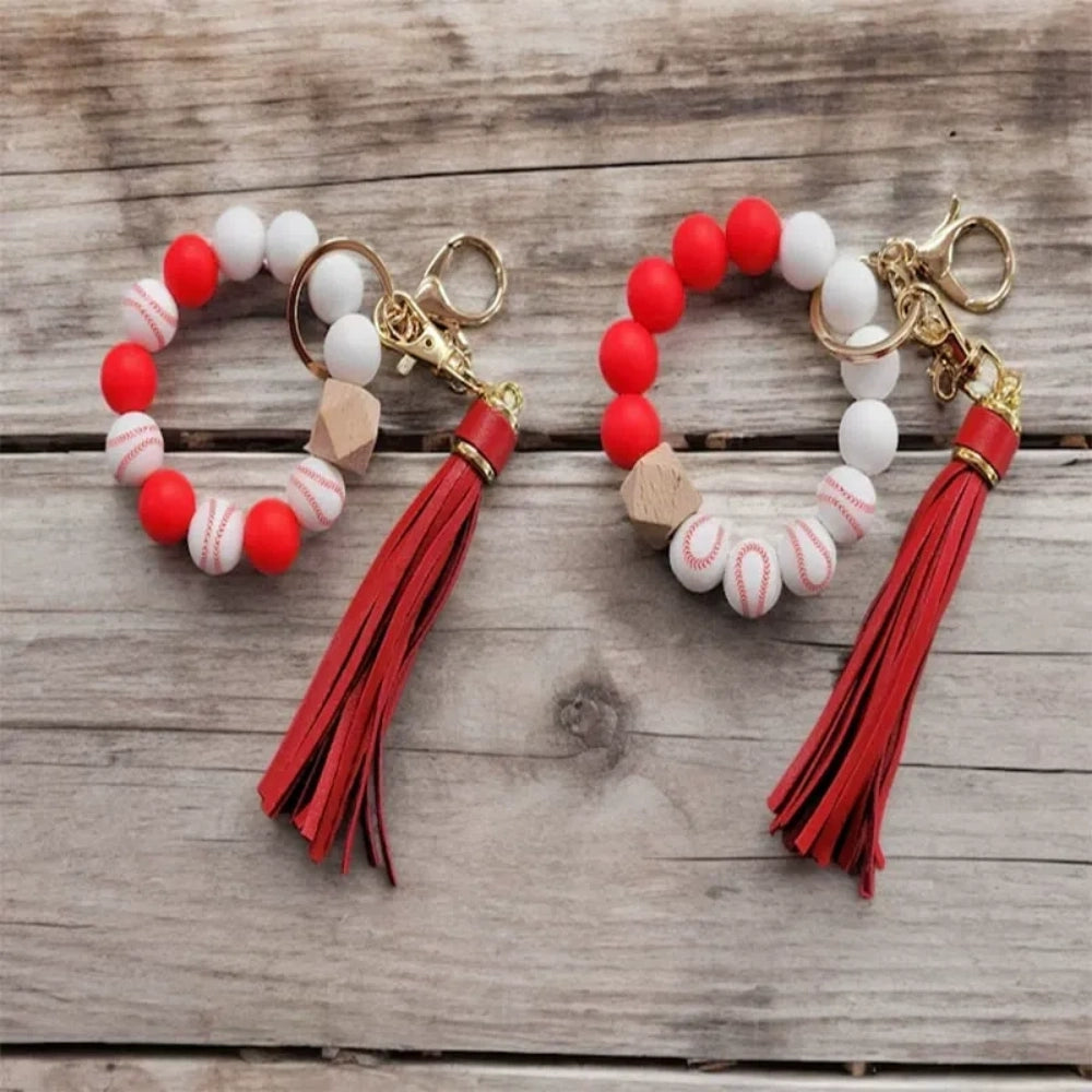 Baseball print silicone beads in red and white shown on beaded wristlet keychains with red leather tassels and gold hardware on a rustic wood background.