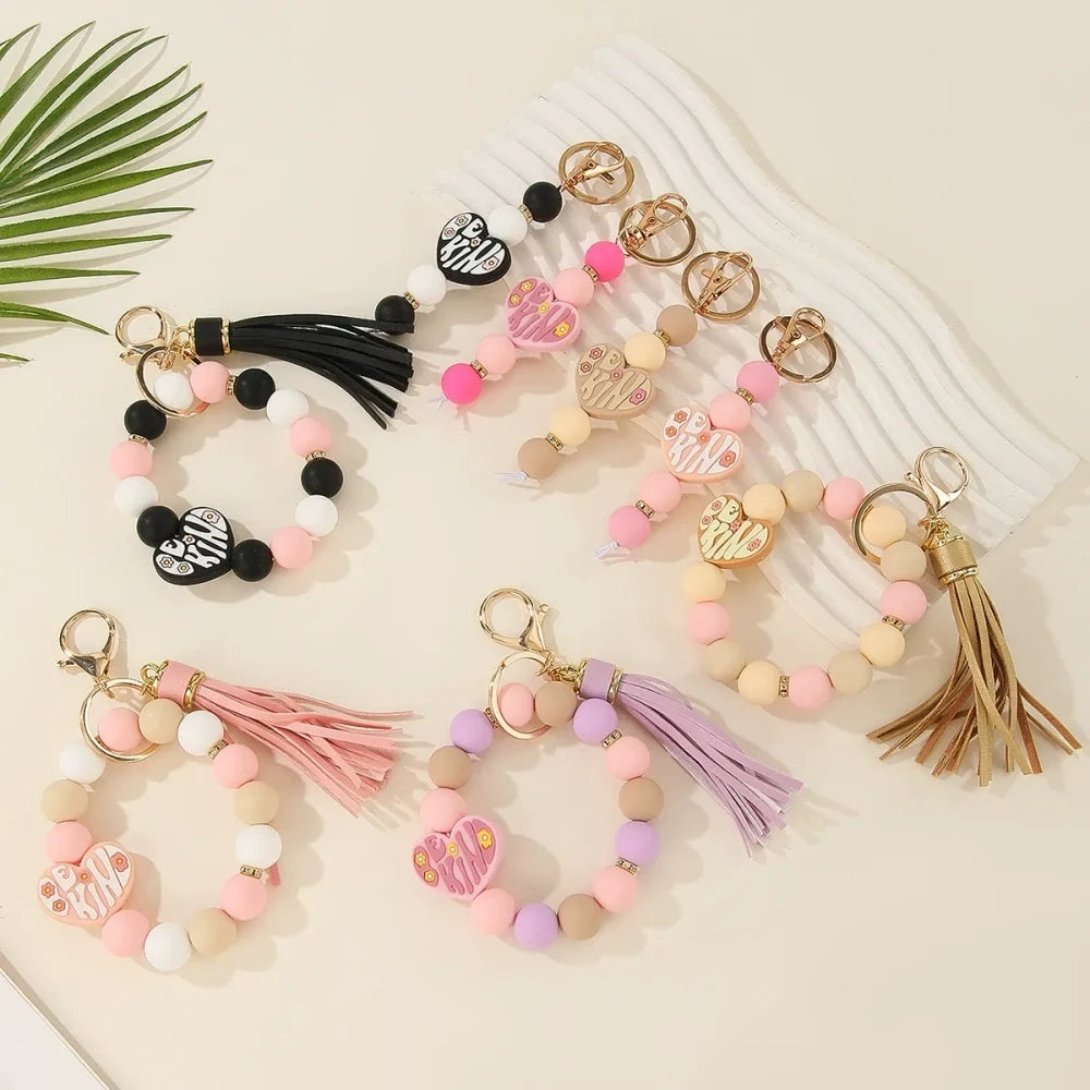Be Kind heart printed silicone beads in pink, black, purple, and neutral tones shown on beaded wristlet keychains with tassels and gold hardware.